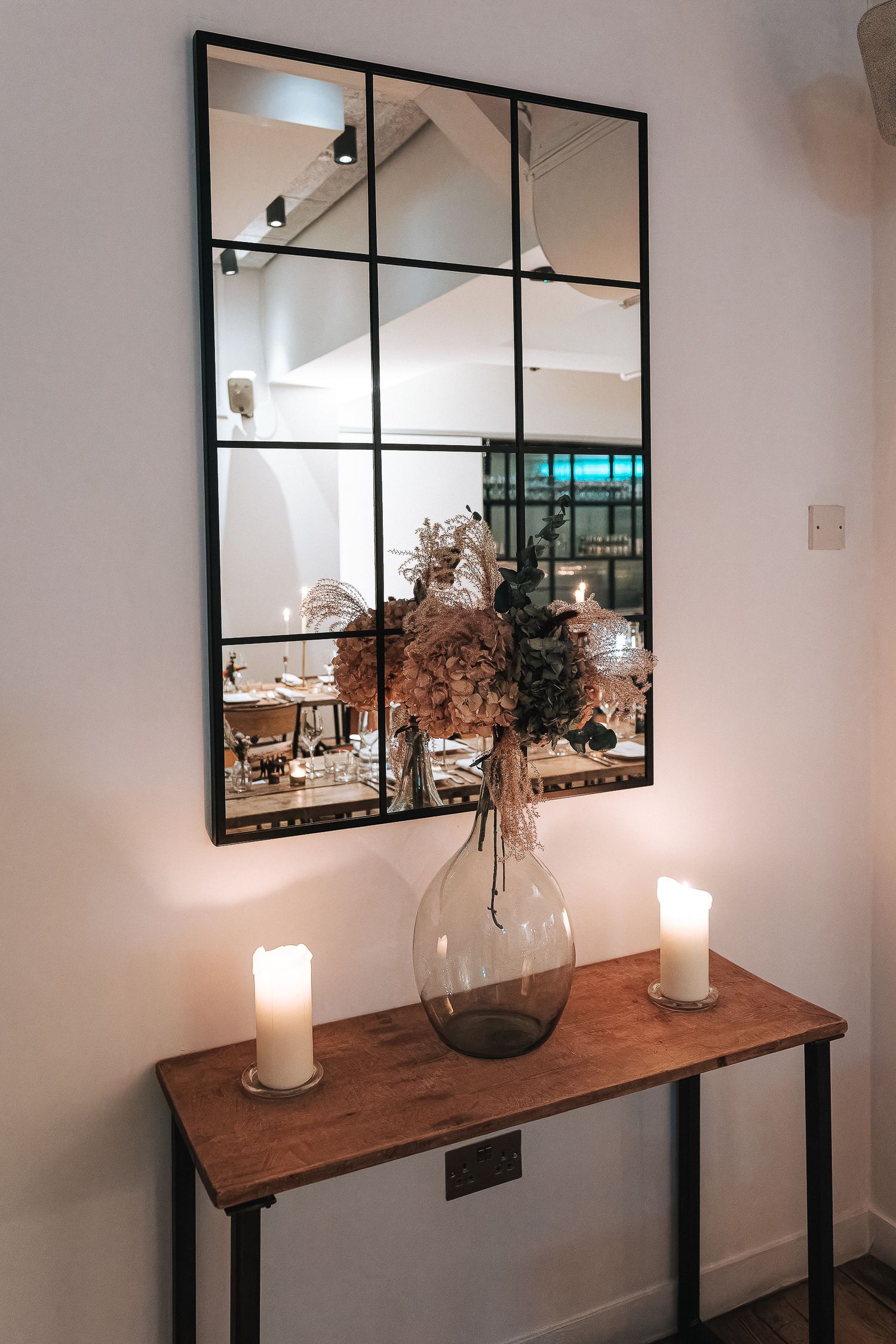 Rustic wooden console table with dried flowers in a vase and black-framed mirrors mounted on a white wall.