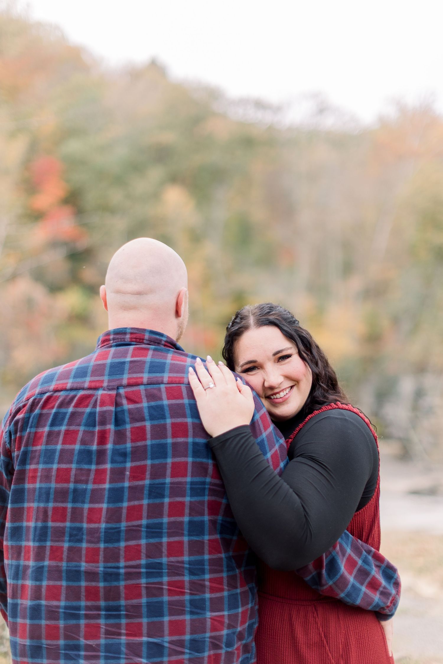 A couple in plaid and burgundy clothing embraces outdoors during an autumn engagement photoshoot.