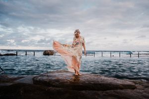 A flowing dress catches the wind on a rocky oceanside pier at sunset.
