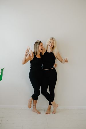Two friends in black outfits posing playfully against white wall making peace signs.