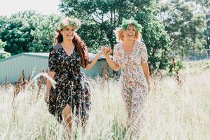 Two friends laughing together while walking through a sunny field of tall grass and wildflowers.