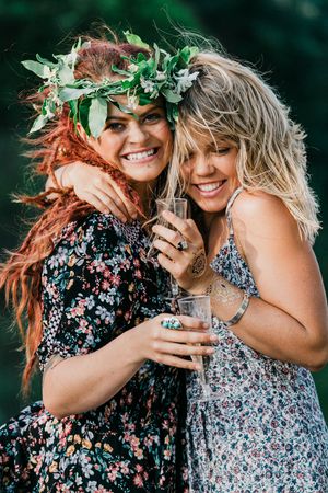 Two friends wearing floral dresses and leaf crowns embrace while laughing outdoors.