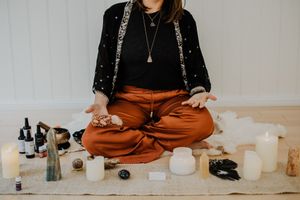 Person in orange pants sits cross-legged with spiritual items and candles on floor.