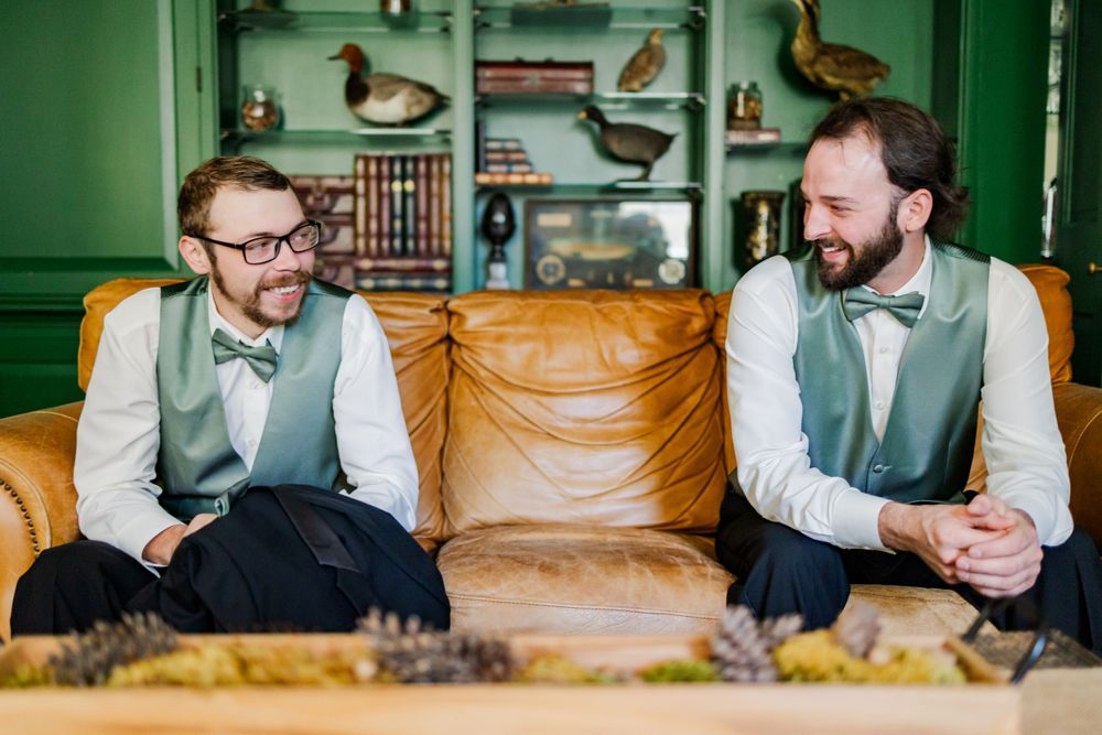 Two groomsmen in matching sage vests share a relaxed moment on vintage leather couch.