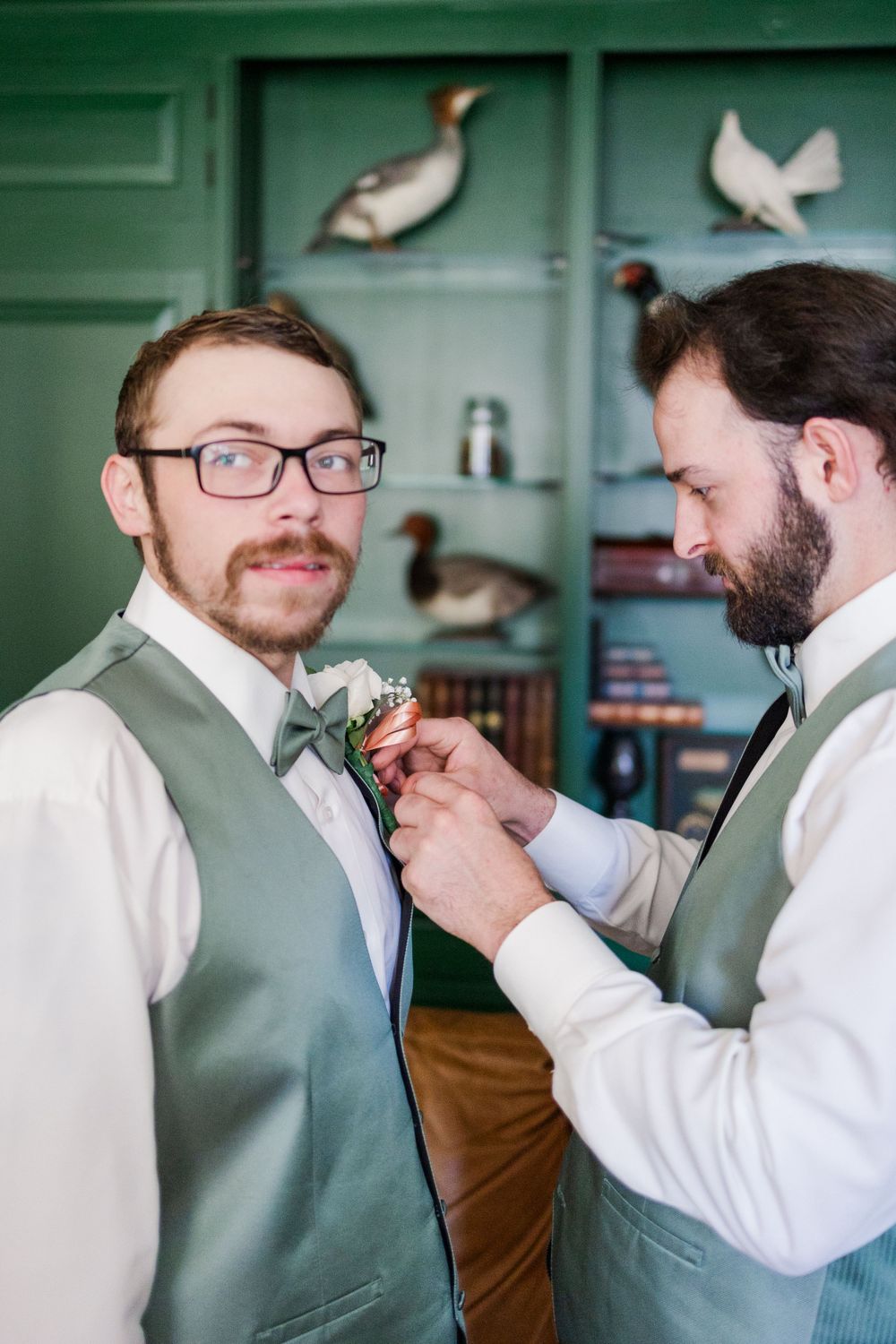 Groomsman helps adjust boutonniere against green wall with duck decor.