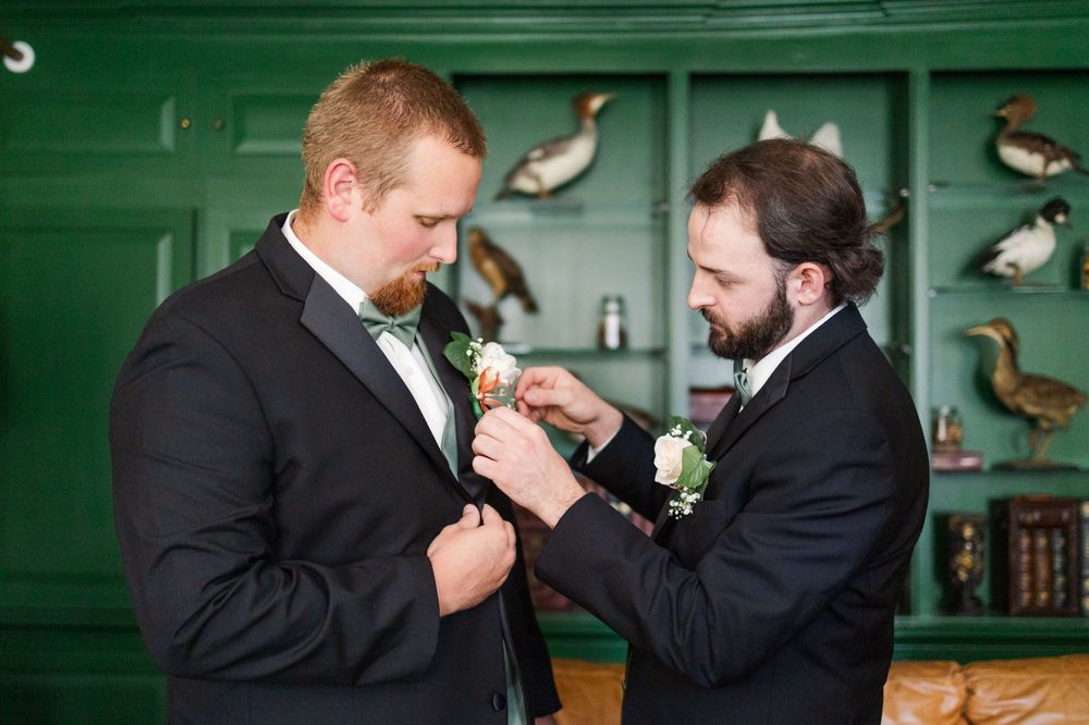 Best man helps secure boutonniere on groom's lapel during wedding preparations.