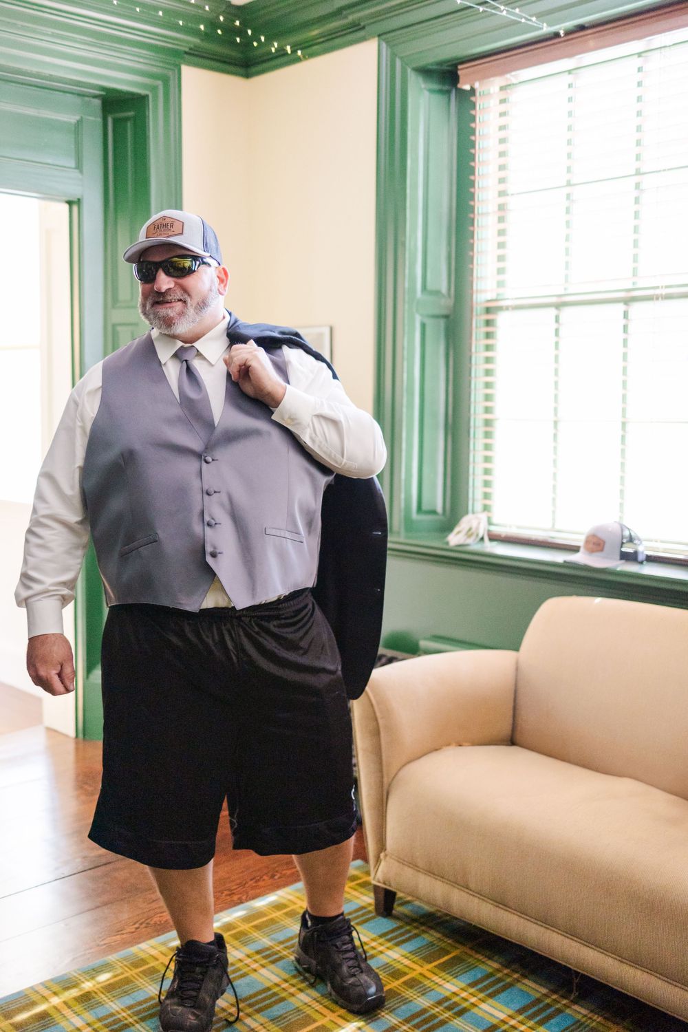 A person in formal attire and a baseball cap stands in a room with green trim and a beige couch.
