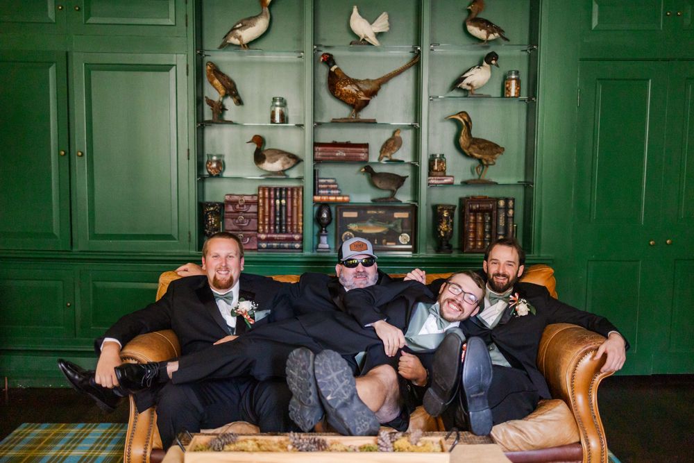 Wedding party lounges casually on a vintage couch against a dark green wall with bird artwork.