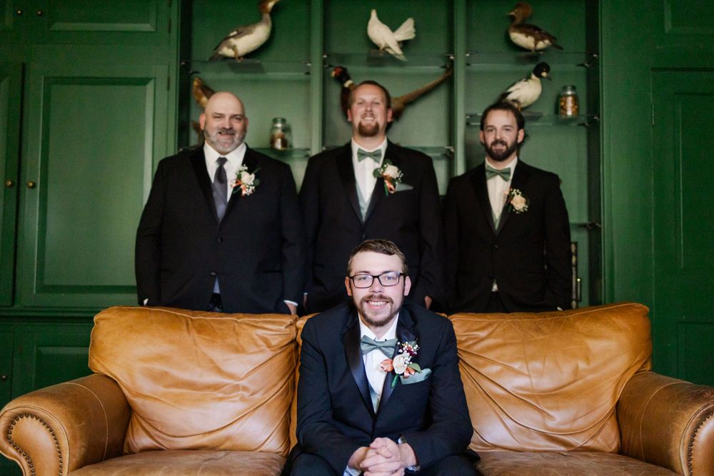 Groomsmen pose for a formal photo on a leather couch against emerald walls with bird decorations.