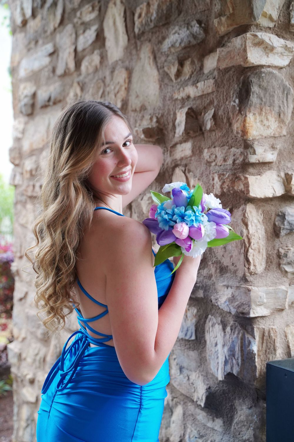 Person in blue formal dress holding colorful floral bouquet while posing against stone wall.