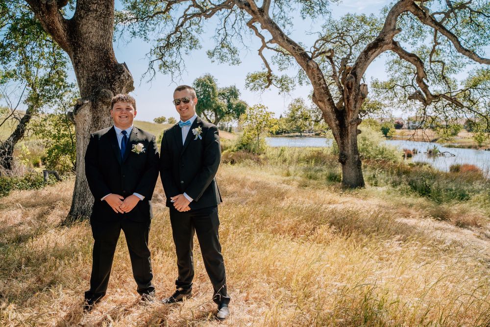Group wearing formal attire stands together on a scenic grassy path under shady trees.