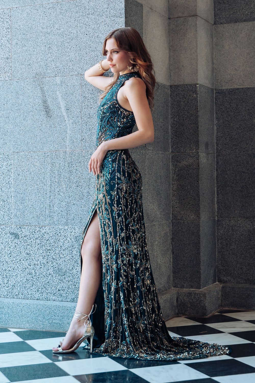 Model posing in elegant teal velvet evening gown against marble pillars.