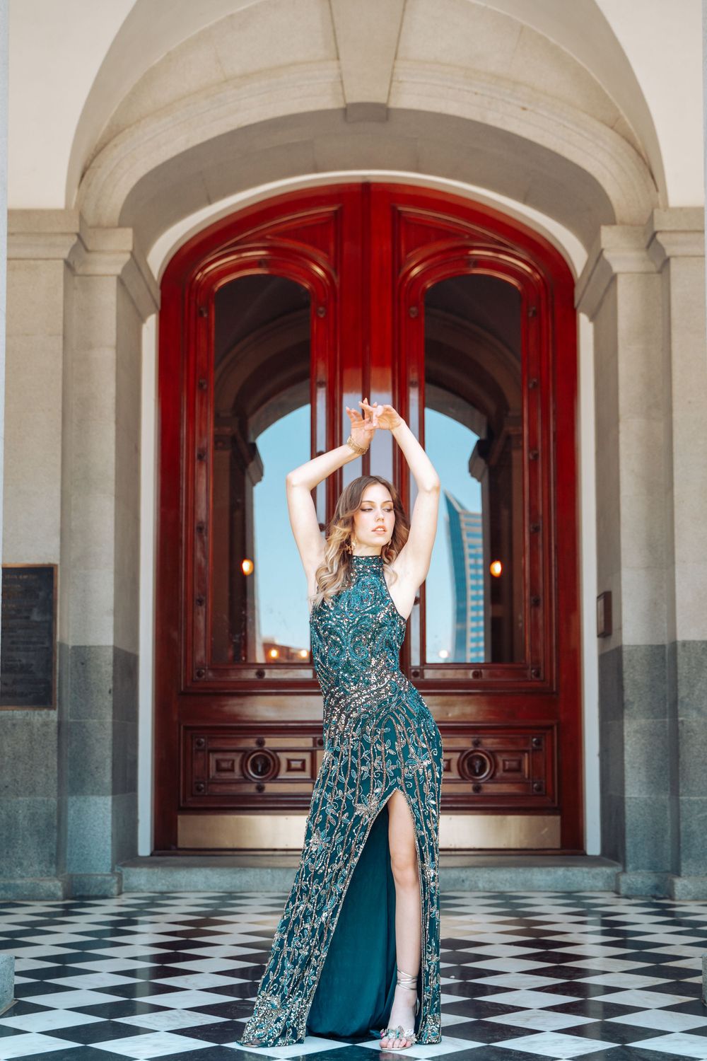 Woman in turquoise patterned dress poses gracefully in classical columned hallway with red arched doors and checkered floor.