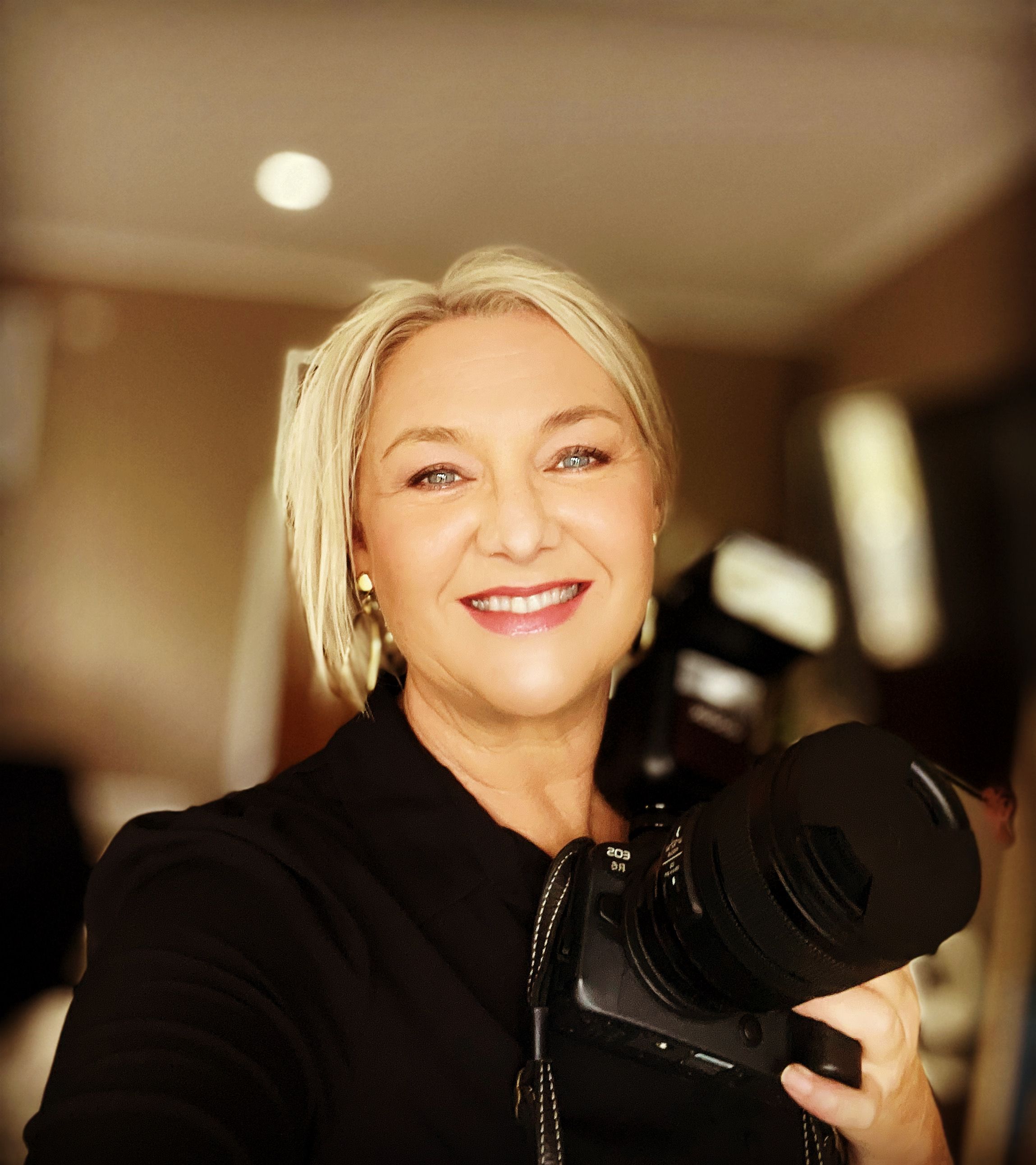 A photographer holding a professional camera while wearing a black top against a warm-toned interior background.