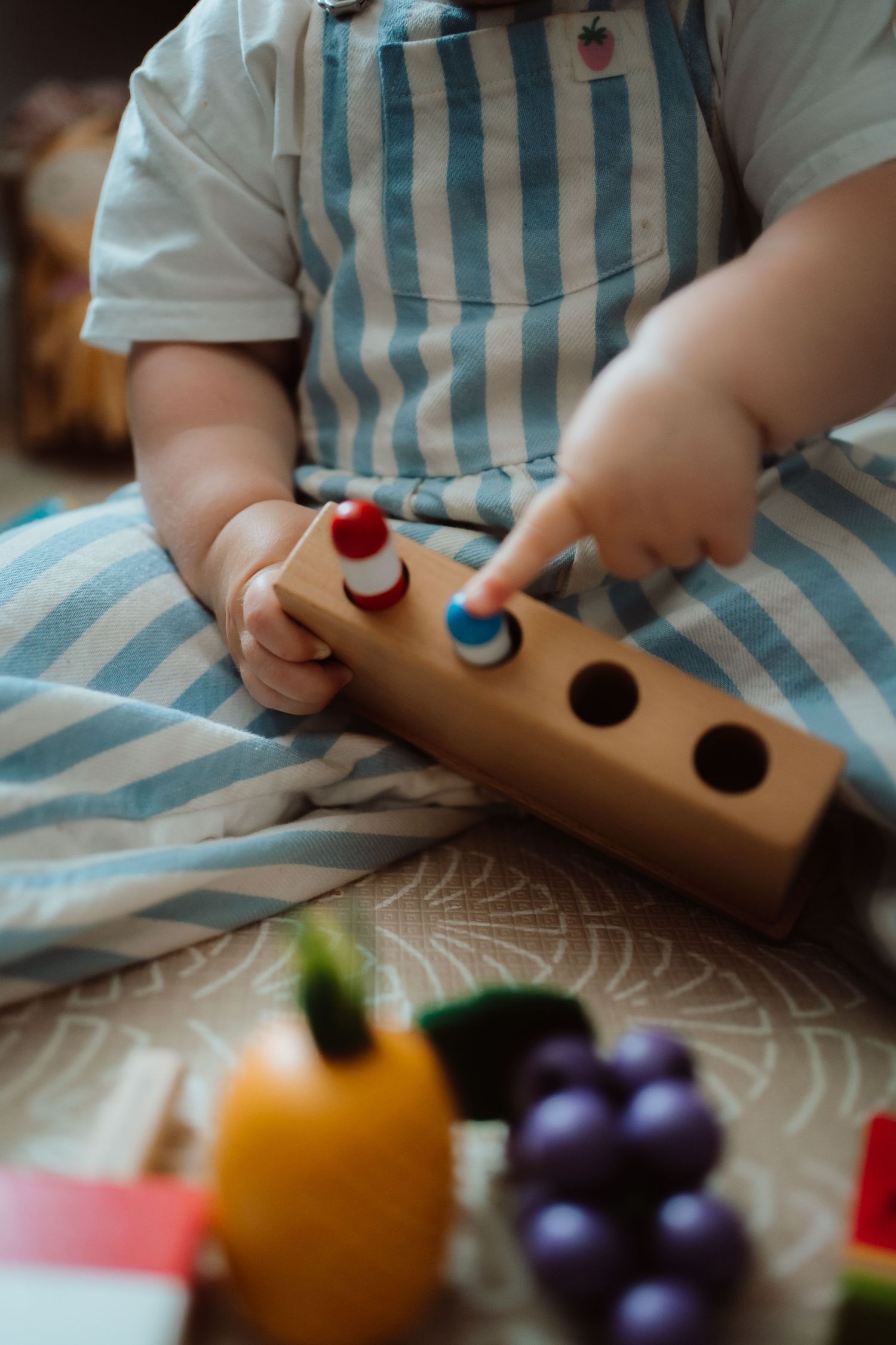 Small hands in blue striped apron play with wooden toy food including colorful grapes and fruit pieces.
