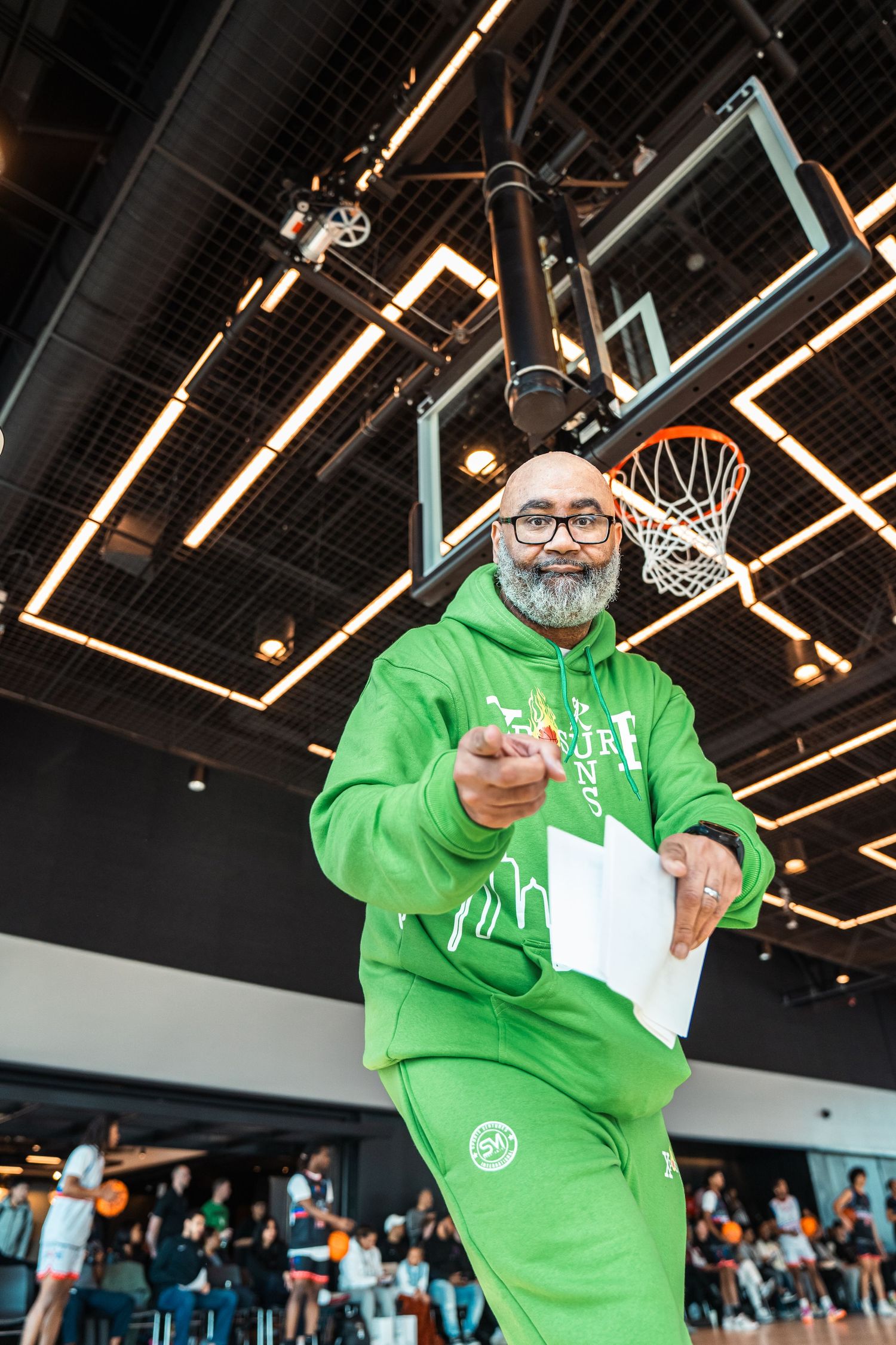 Man in bright green hoodie standing in front of basketball court ceiling and scoreboard.