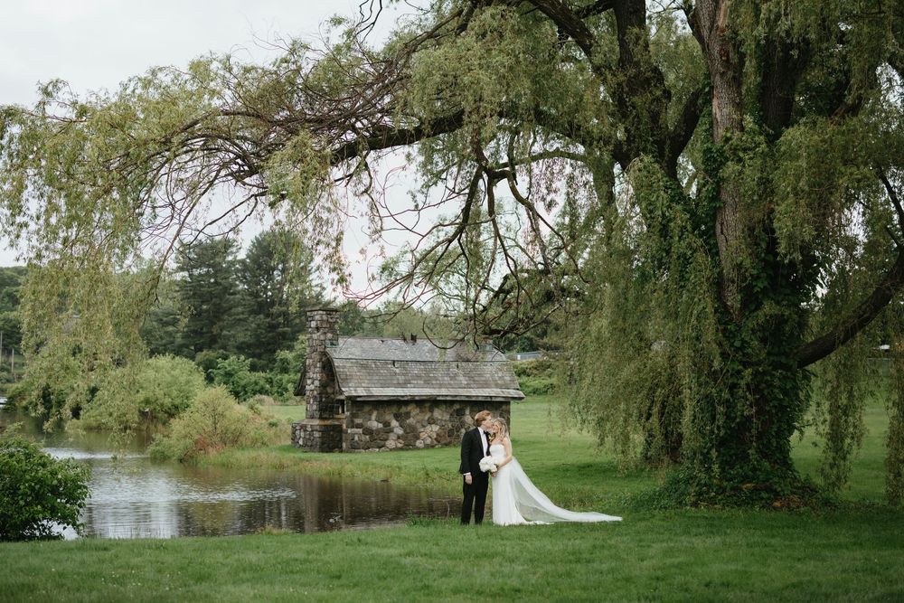 A couple poses beneath drooping willow trees near a tranquil pond in a romantic garden setting.