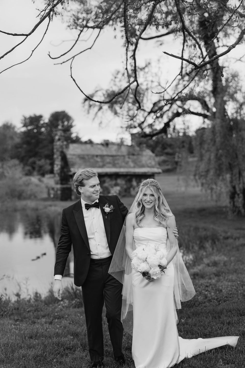 A bride and groom pose together in formal wedding attire near a lake during an outdoor photo session.