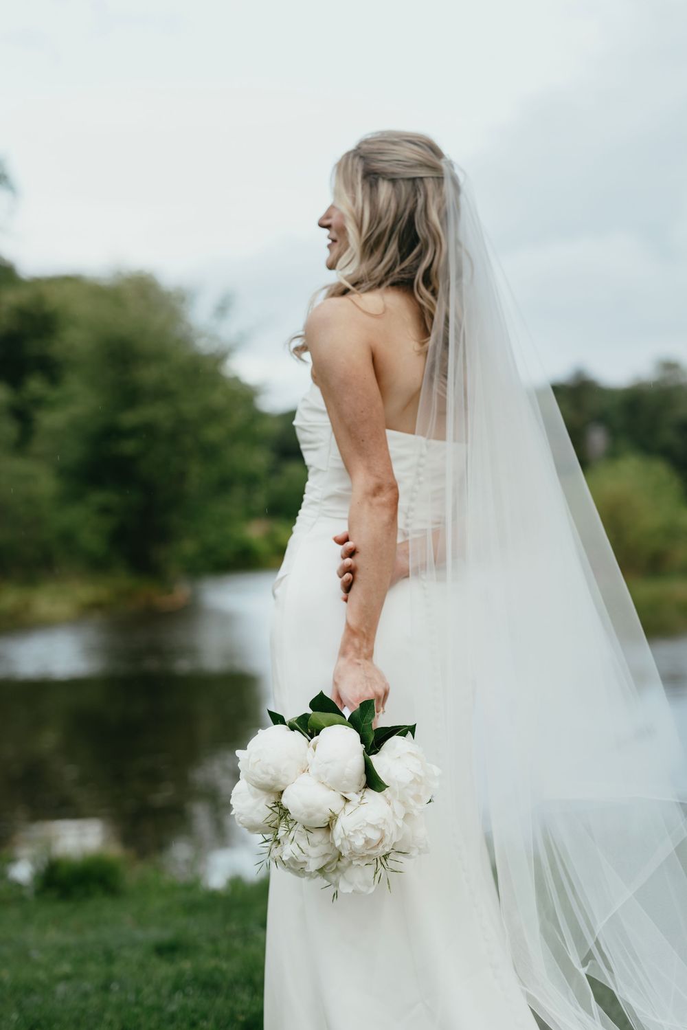 An elegant bride in a white wedding gown holds a bouquet of white flowers against a natural outdoor backdrop.
