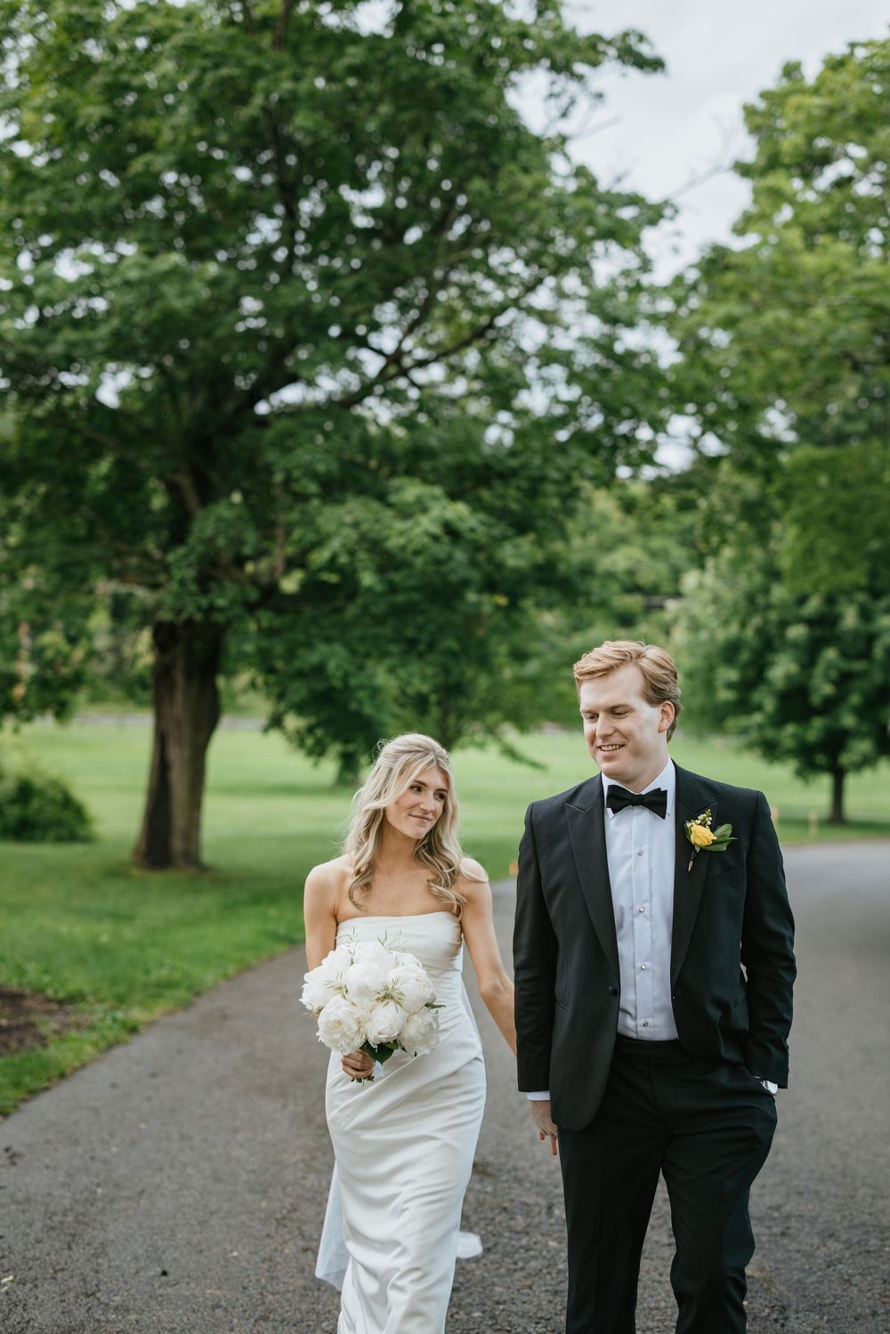 A couple in formal wedding attire walks together on a tree-lined path on their special day.