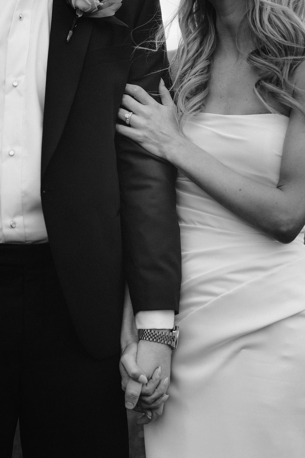 Close-up details of a bride and groom holding hands, showing formal wedding attire in black and white.