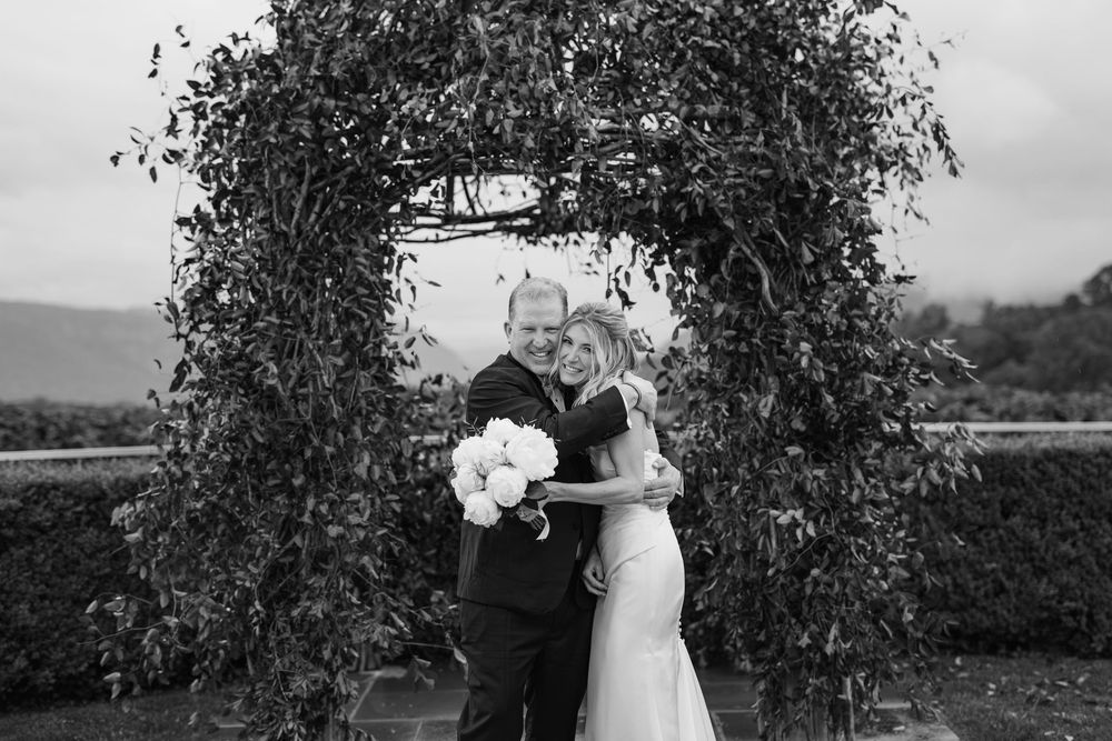 A black and white wedding photo showing a couple embracing under a floral arch with mountains in the background.
