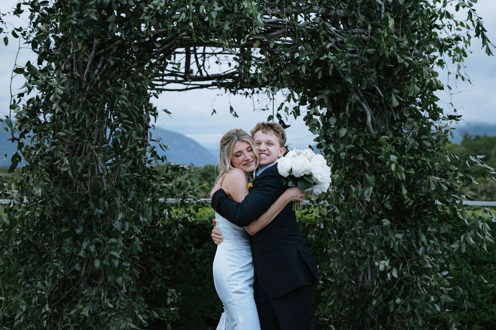 A couple embraces while standing in front of lush green trees and foliage on a sunny day.