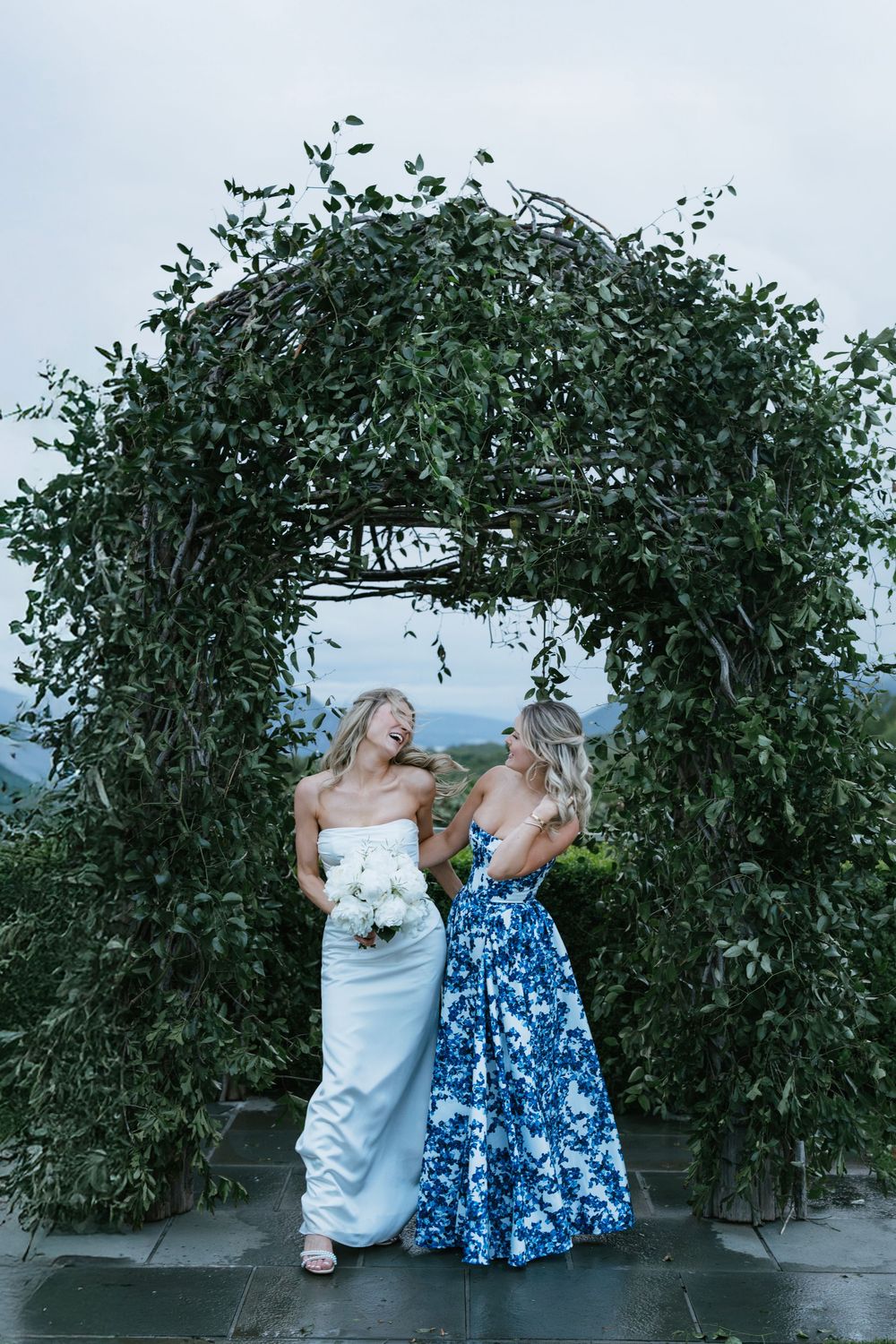 Two women in formal dresses share a joyful moment under a lush green garden archway against a cloudy sky.