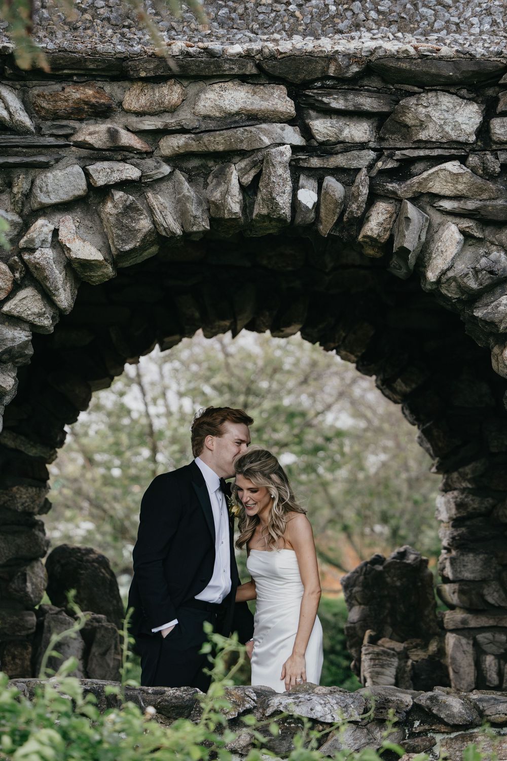 A romantic couple shares intimate moments through stone archways in a garden setting during their wedding photoshoot.