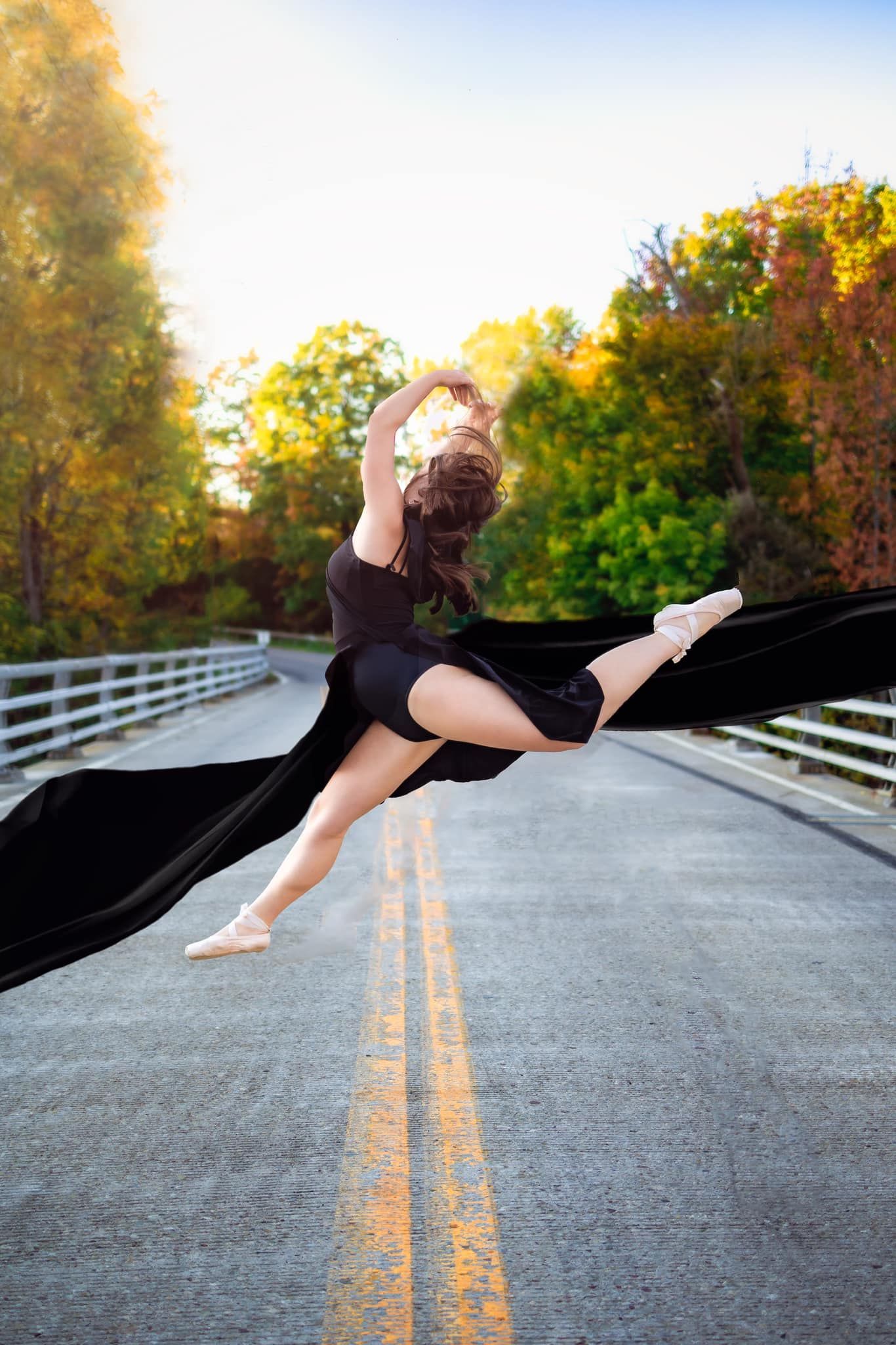 Graceful dancer in black performing a dramatic leap on a road with autumn trees in background.