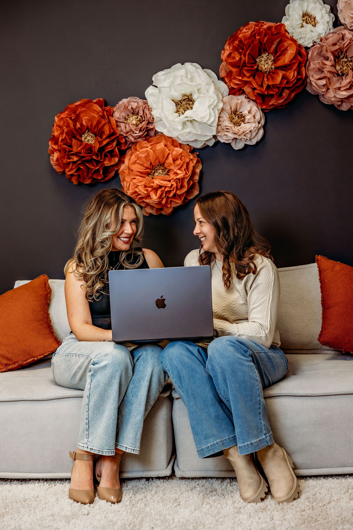Two people sharing laptop on couch beneath decorative floral wall display.