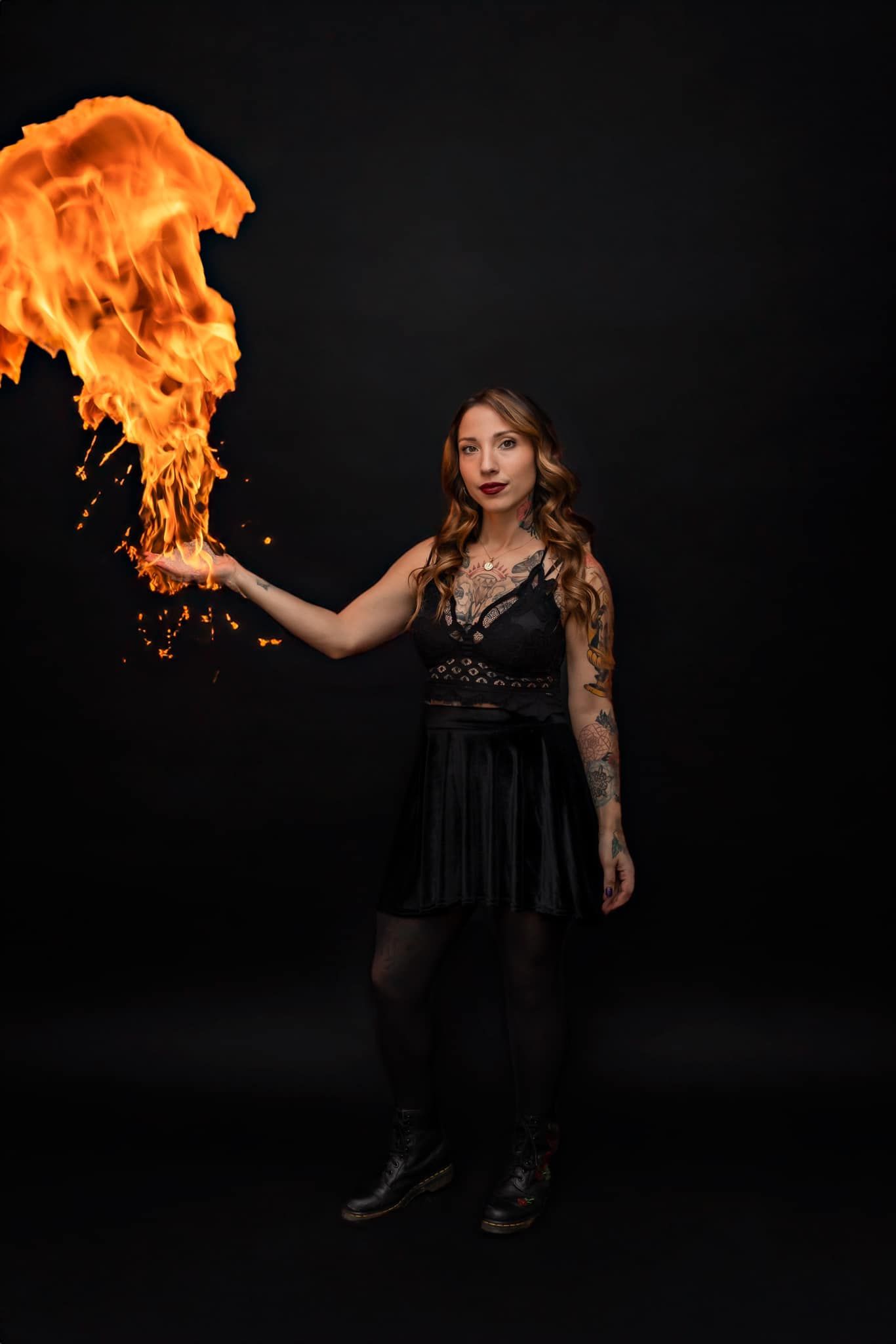 Figure in black dress holding dramatic orange flame against dark background.