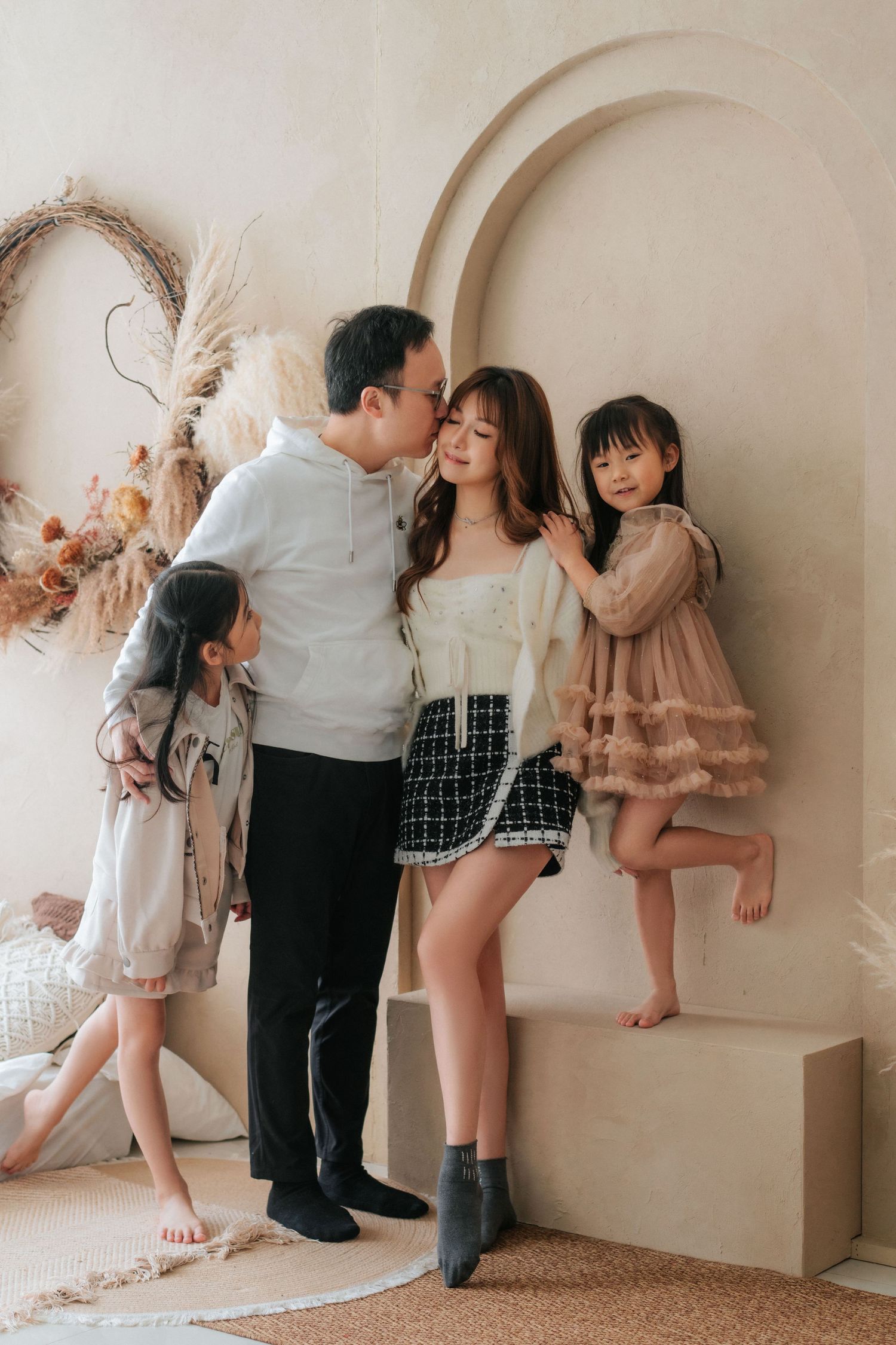 Asian family portrait with parents and two daughters posing in a neutral-toned studio with dried floral decor.亞洲家庭合影，父母和兩個女兒在以乾燥花裝飾的中性色調攝影棚中