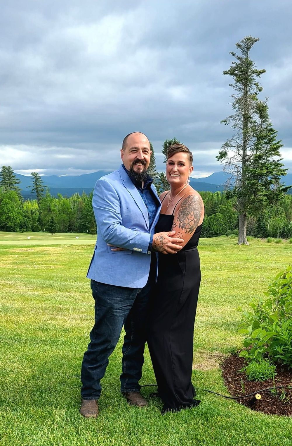 Couple embraces on scenic mountain overlook with evergreen trees and cloudy sky in background.