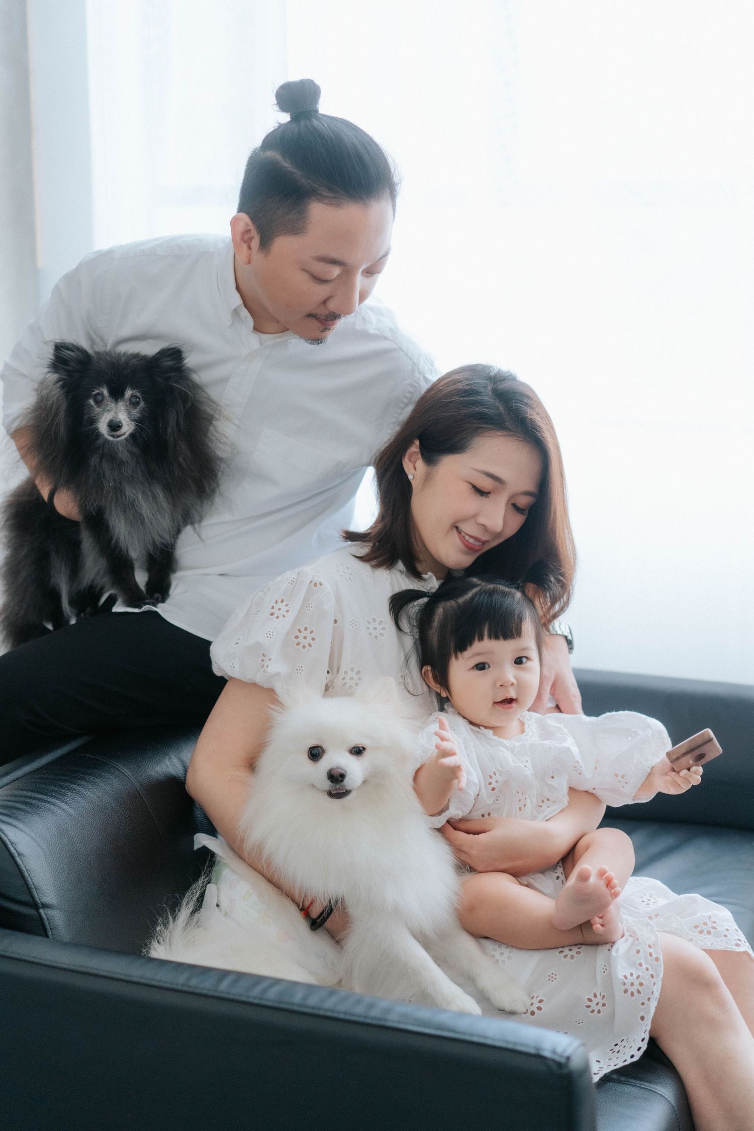 Asian family with two Pomeranian dogs sitting on black leather sofa, parents smiling at baby girl holding smartphone.一個亞洲家庭帶著兩隻博美犬坐在黑色皮沙發上