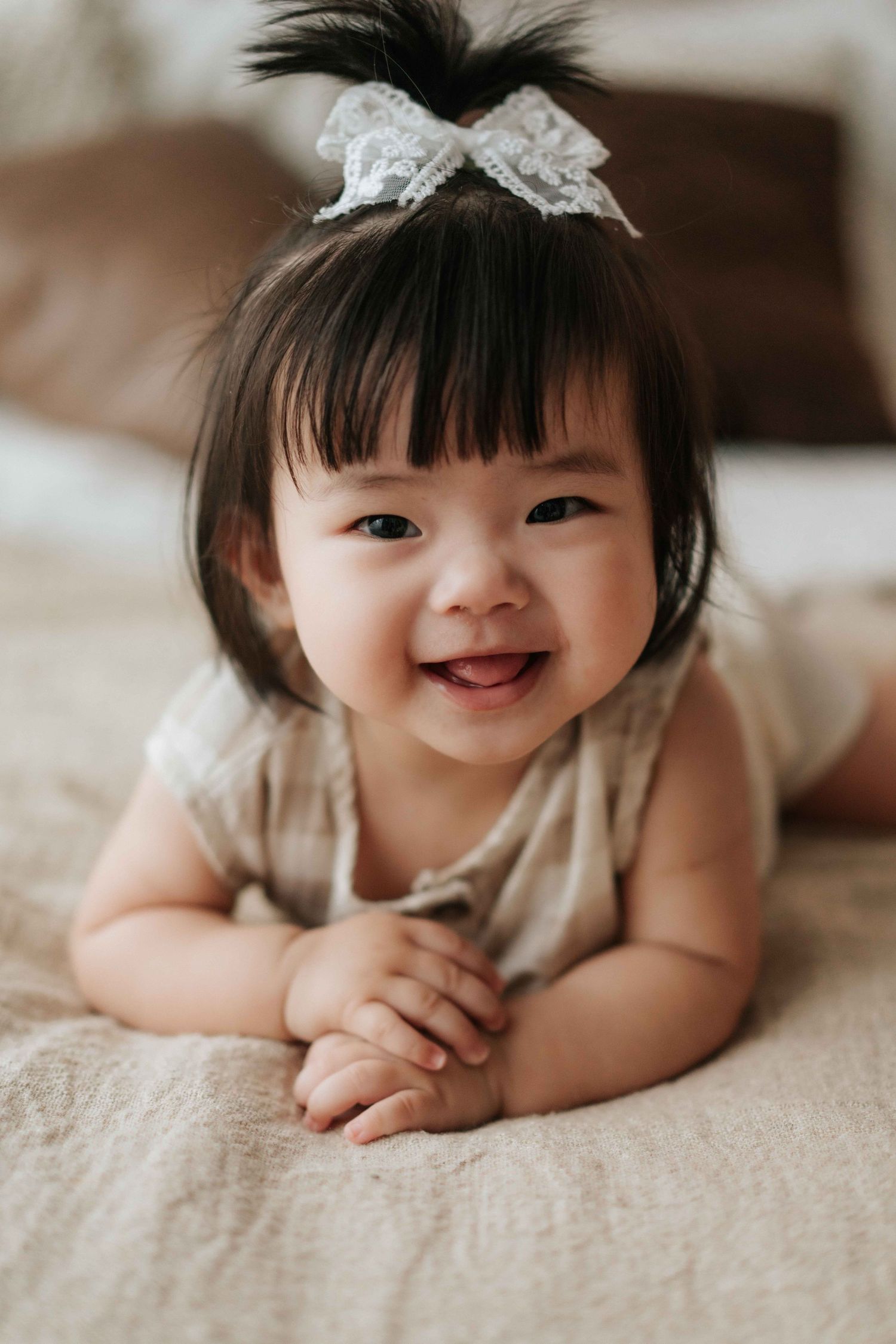 Adorable smiling Asian baby girl with dark hair and white lace bow lying on bed wearing beige onesie.可愛的亞洲女嬰，有著深色的頭髮和白色蕾絲蝴蝶結，穿著米色連身衣，躺在床上微笑。