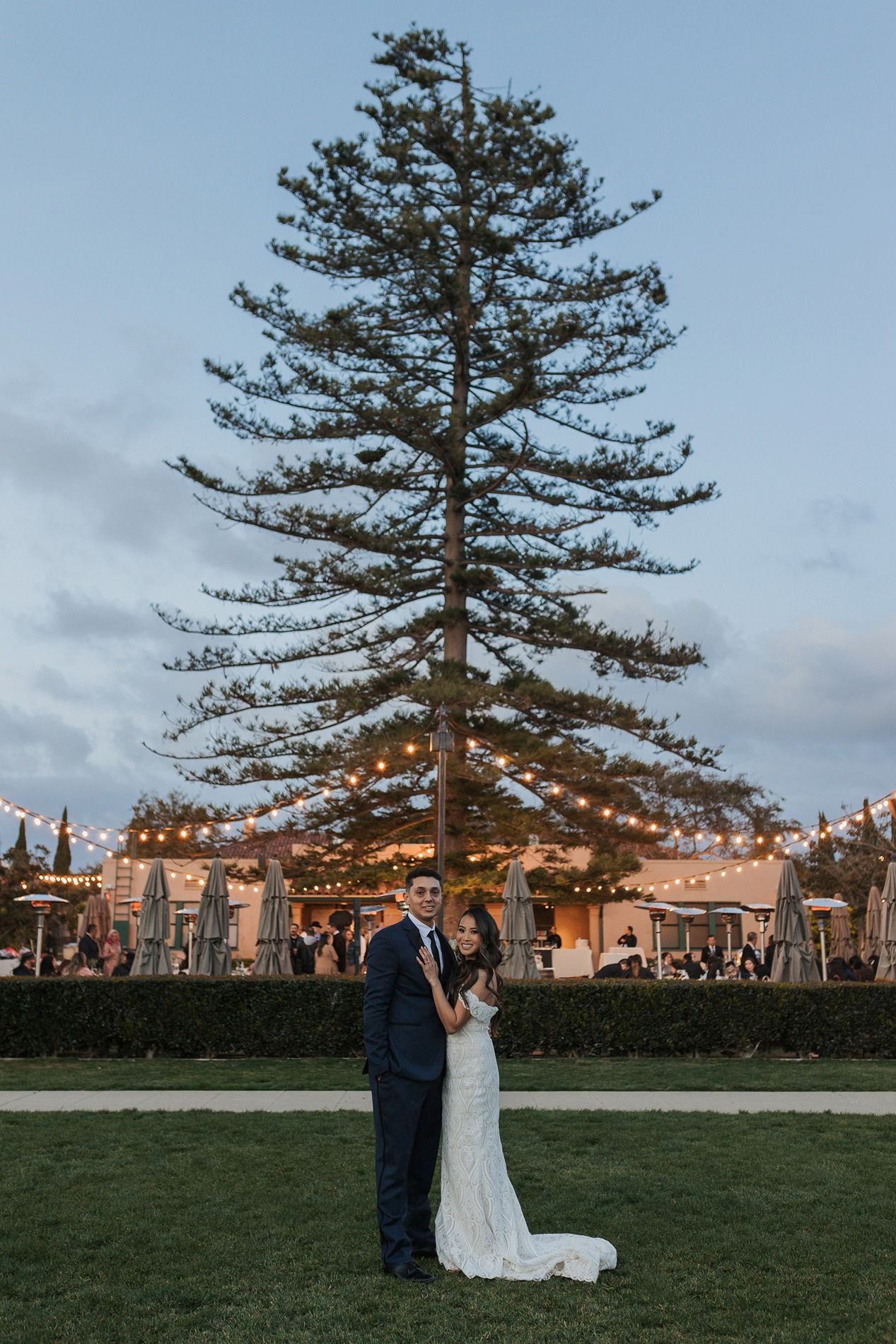 A couple in wedding attire stands beneath a towering pine tree on a manicured lawn at sunset at Stone Brewing in San Diego.