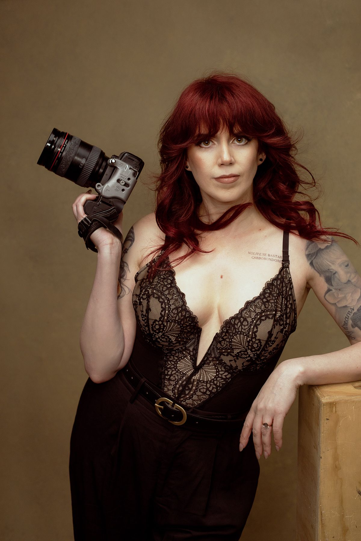 Professional photographer in black lace bodysuit and high-waisted pants poses with DSLR camera against neutral backdrop.