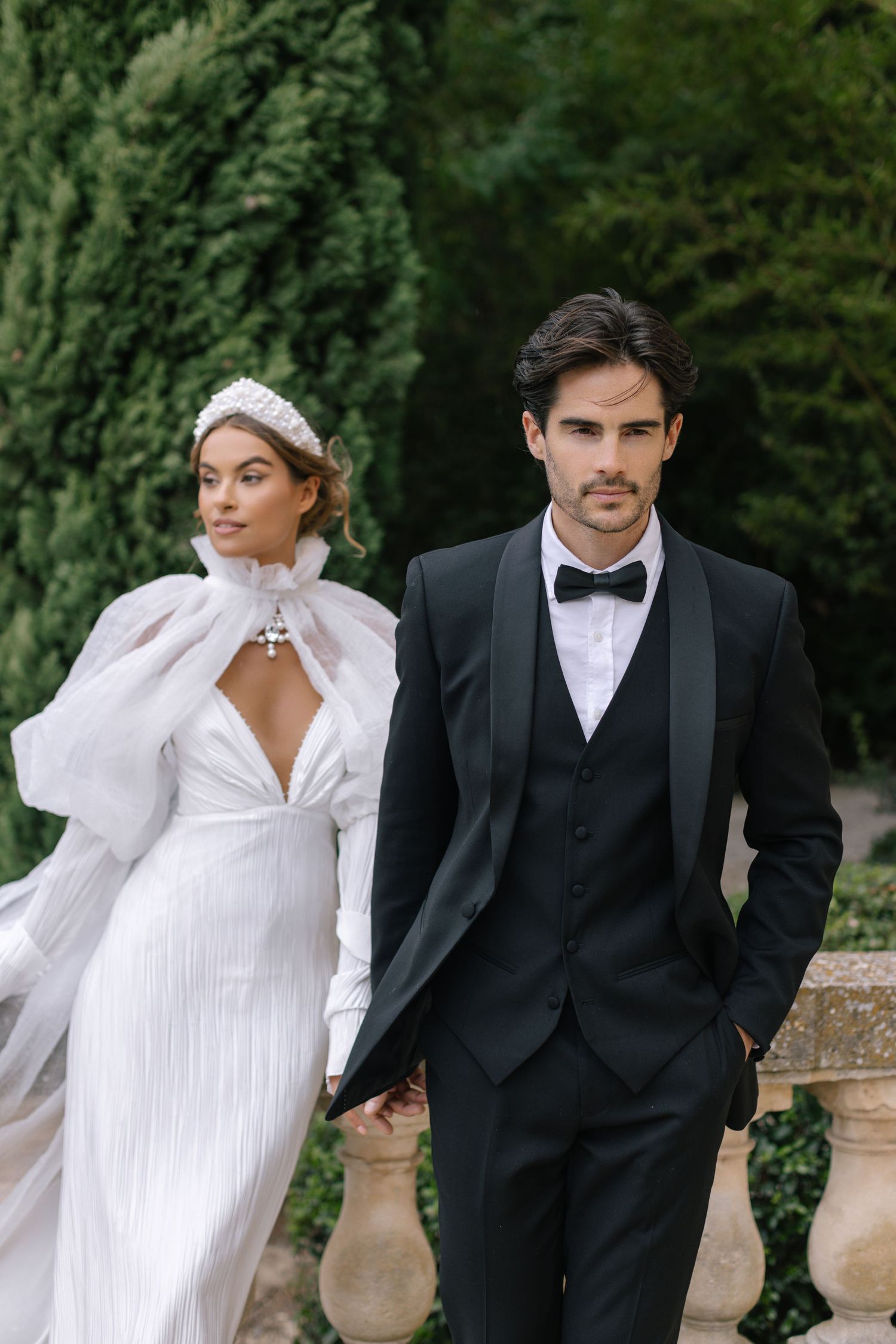 A couple walks down an outdoor aisle lined with flowers and cypress trees leading to an elegant villa entrance.