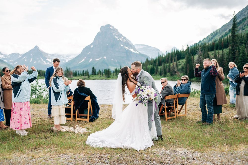 A scenic mountain wedding celebration takes place in a meadow with dramatic peaks and evergreen forests in the background.