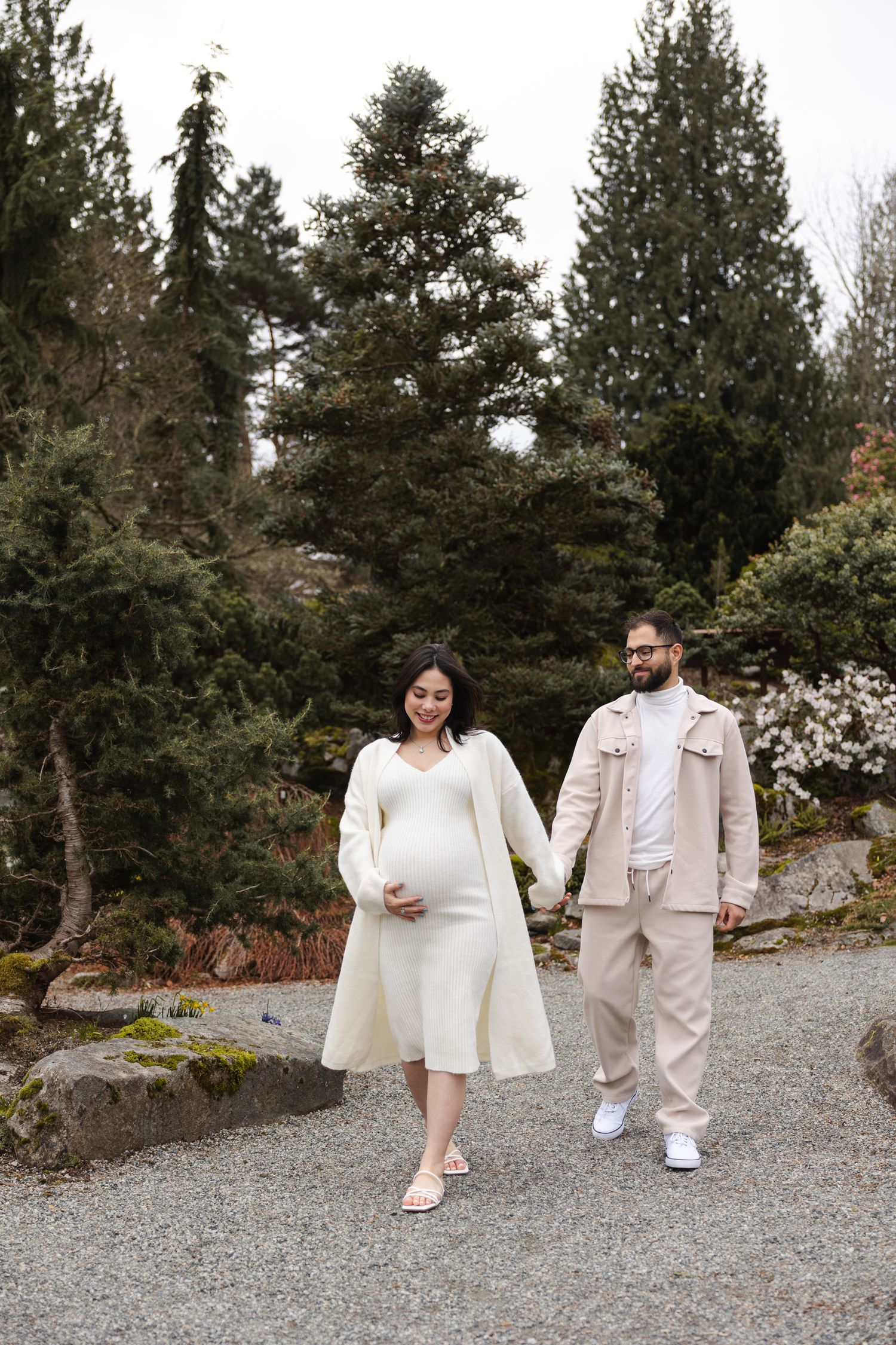 Couple in matching white outfits pose on a gravel path surrounded by evergreen trees.