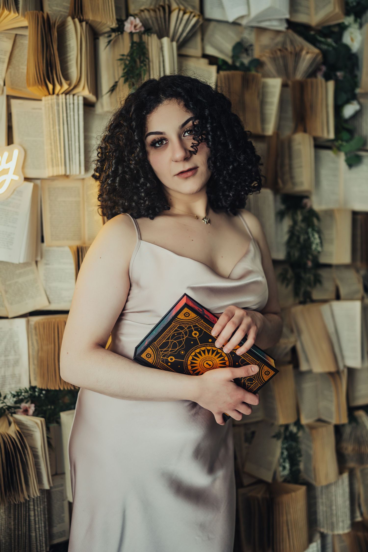 A curly-haired woman in a satin slip dress holds an ornate celestial book while standing before a wall of open books.