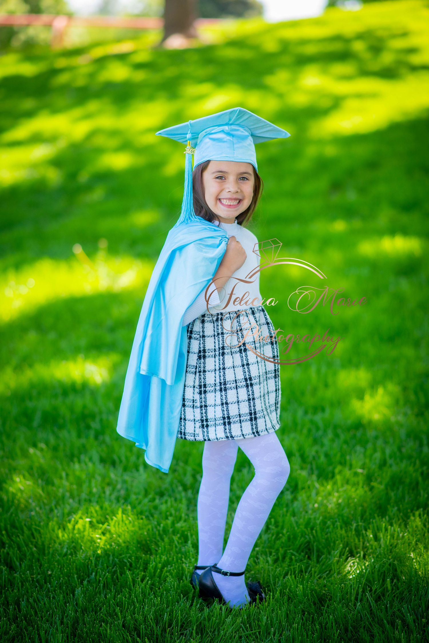 A child in a light blue graduation cap and gown poses on grass on a sunny day.