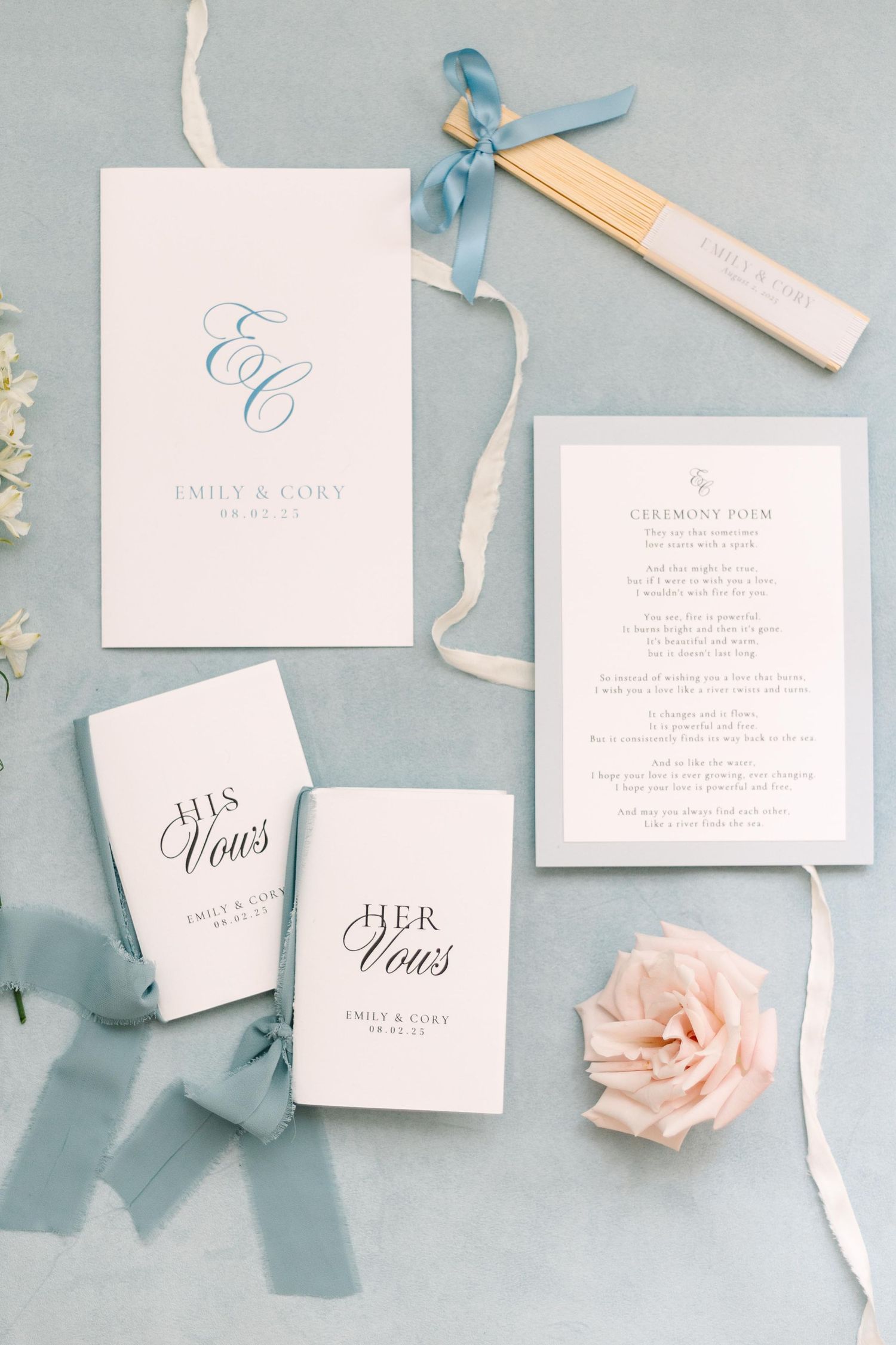 Elegant wedding stationery flatlay with blush pink roses and dusty blue ribbons on light gray background.
