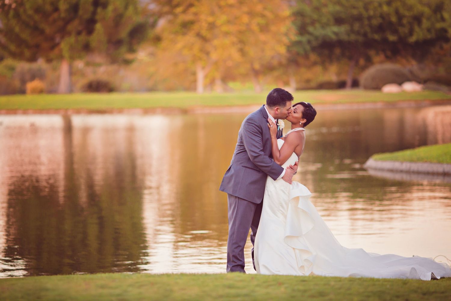 A bride and groom share a romantic kiss by a serene pond, surrounded by lush greenery during their outdoor wedding at Anthem Country Club.