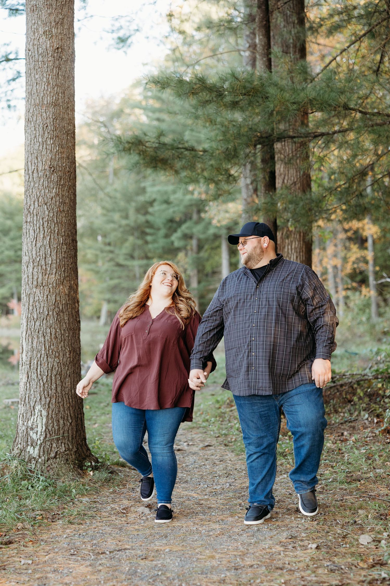 A couple walks hand in hand along a wooded forest trail surrounded by tall pine trees on an autumn day.