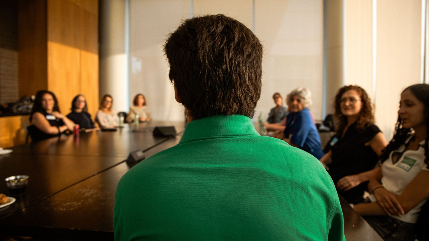 Person in green shirt seen from behind during a business meeting with diverse attendees seated around conference table.