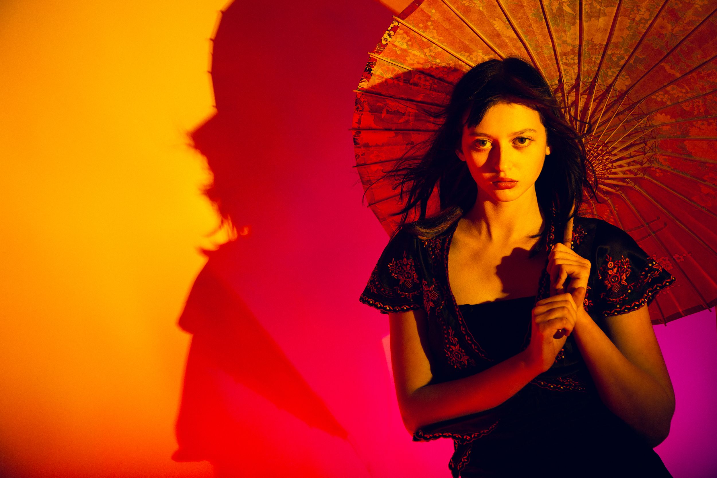 A person in dramatic lighting poses with a red paper umbrella against vibrant orange and pink background.