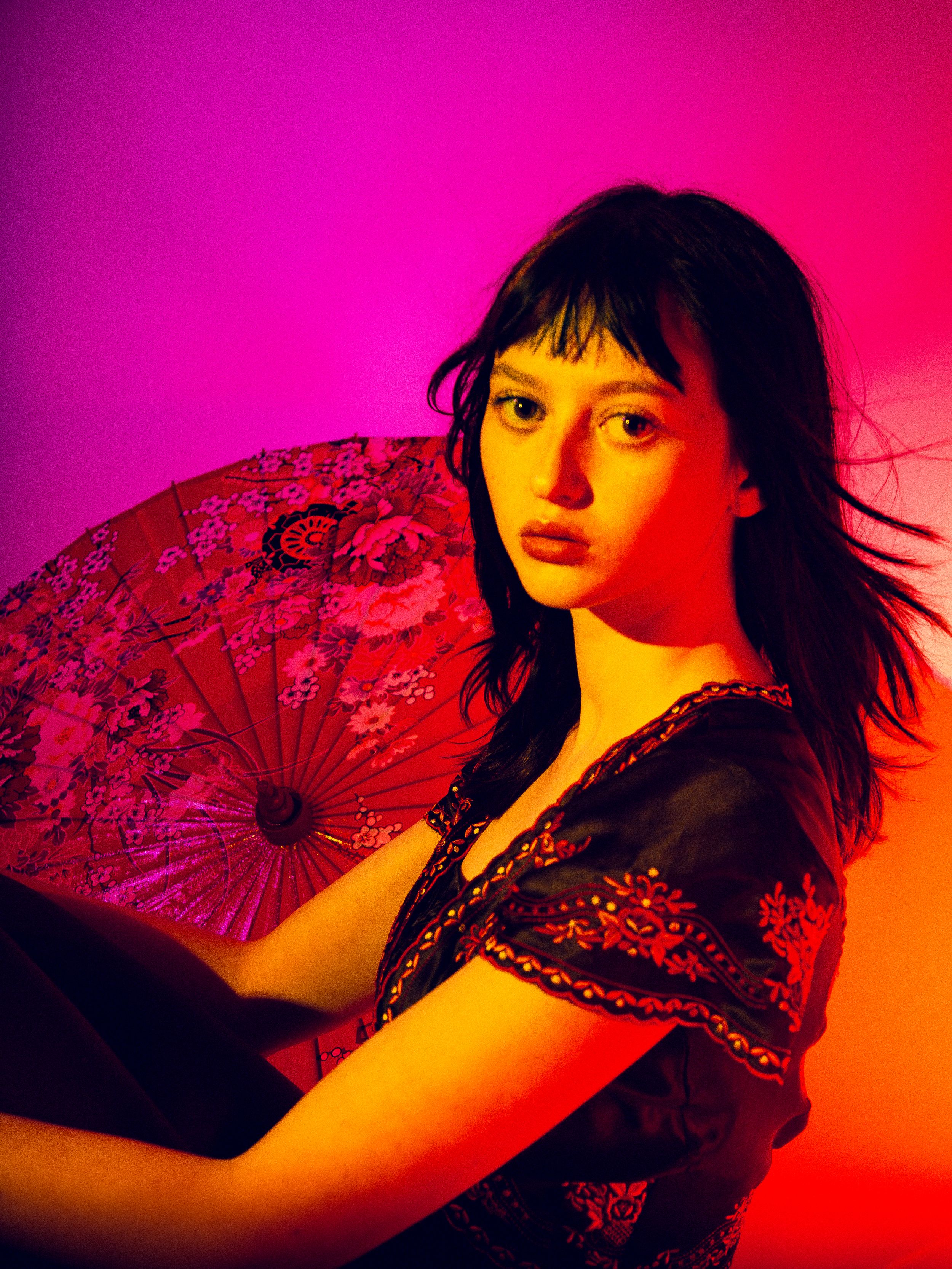 Artistic portrait with red fan against vibrant pink and orange gradient background in dramatic lighting.