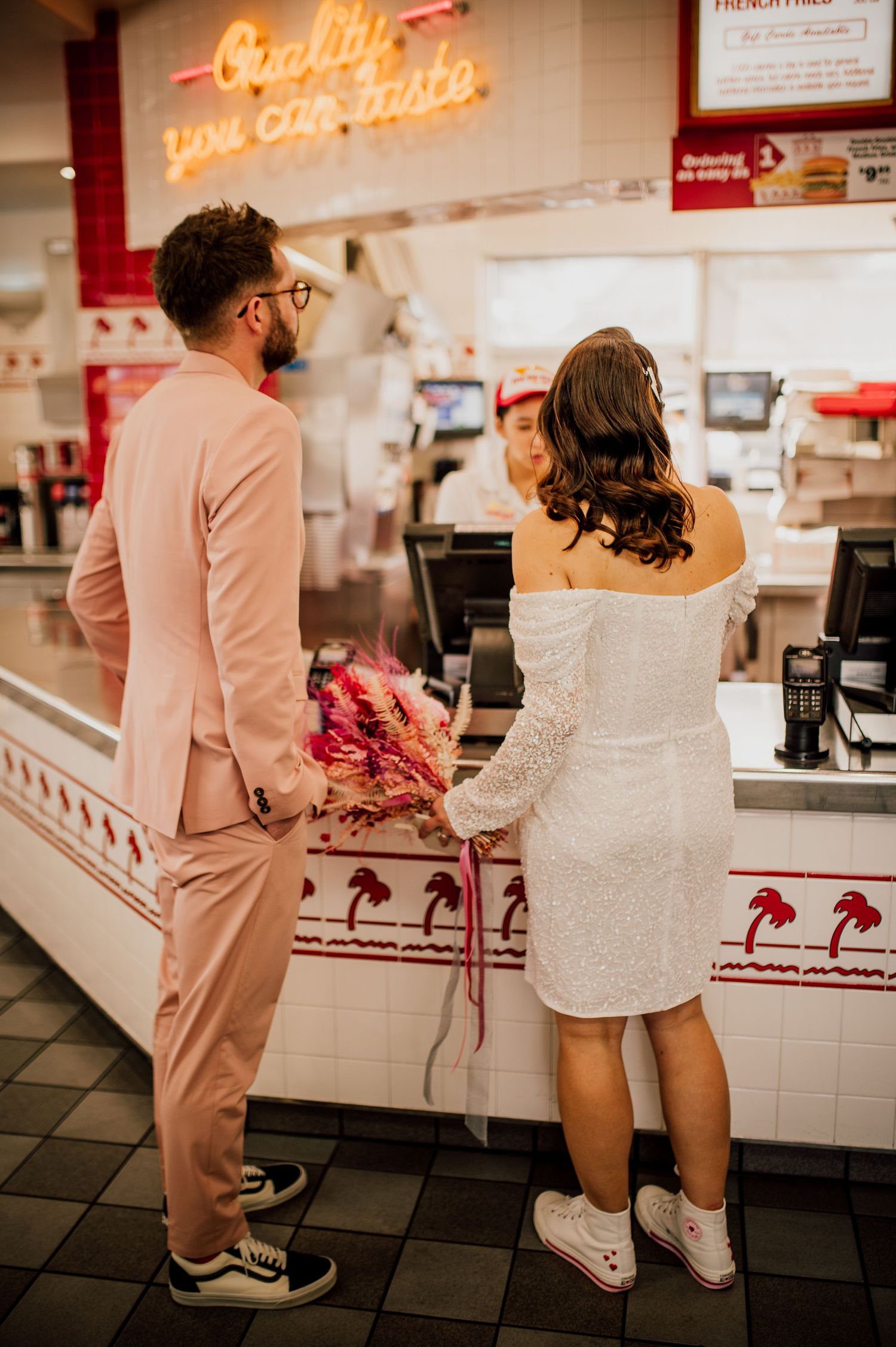 A couple in formal attire orders at a Chick-fil-A counter, with the woman wearing white and the man in a pink suit.